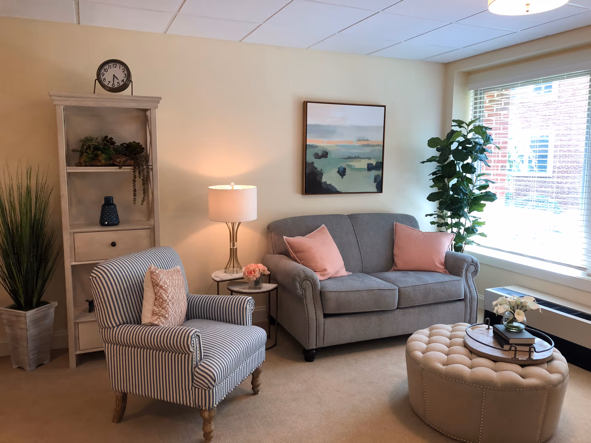 A cozy living room with a gray loveseat adorned with two pink pillows, a striped armchair with a pink cushion, a round tufted ottoman with a tray holding a small vase of white flowers and books, a tall potted plant near a large window with blinds, a floor lamp on a small side table, and a tall shelf with decorative items and a clock on top.