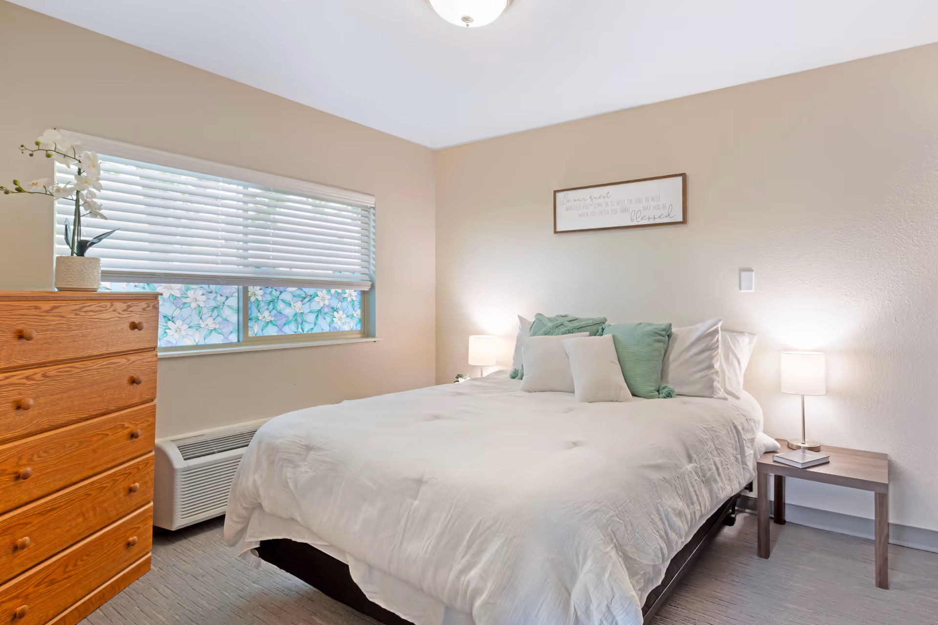 Bright, simply furnished bedroom with a bed, bedside tables and lamps, a wooden dresser, and a window with blinds.