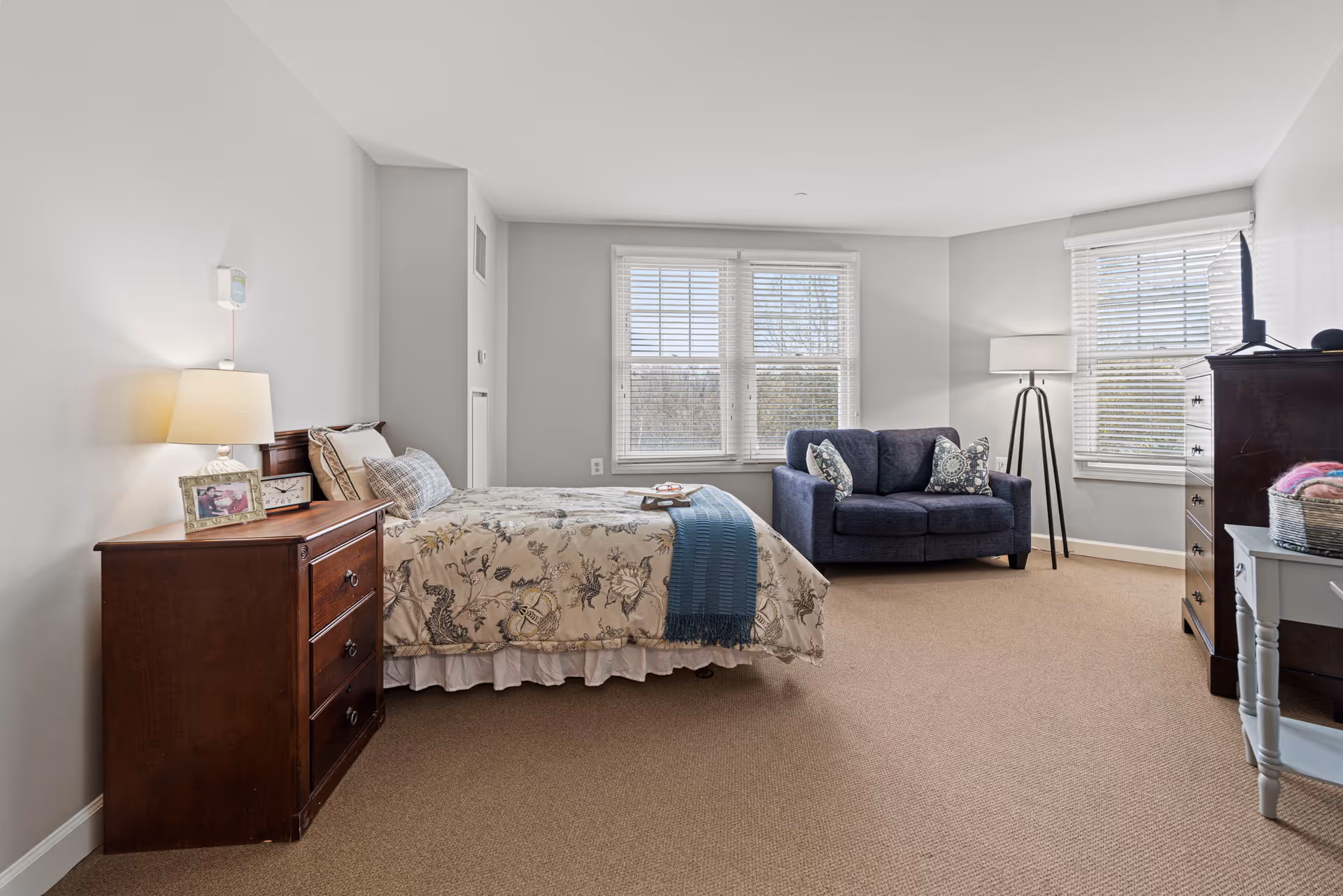 Bright, neatly furnished bedroom with a bed, wooden nightstand, blue loveseat, and windows with blinds.