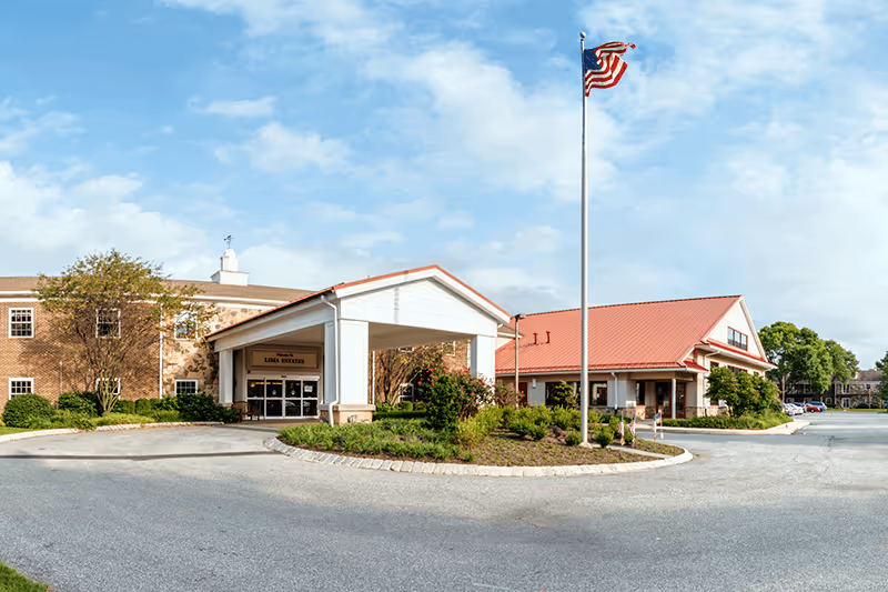 Exterior view of Lima Estates senior living facility showing a two-story brick building with a covered entrance, landscaped greenery, and an American flag on a flagpole in front under a partly cloudy sky.