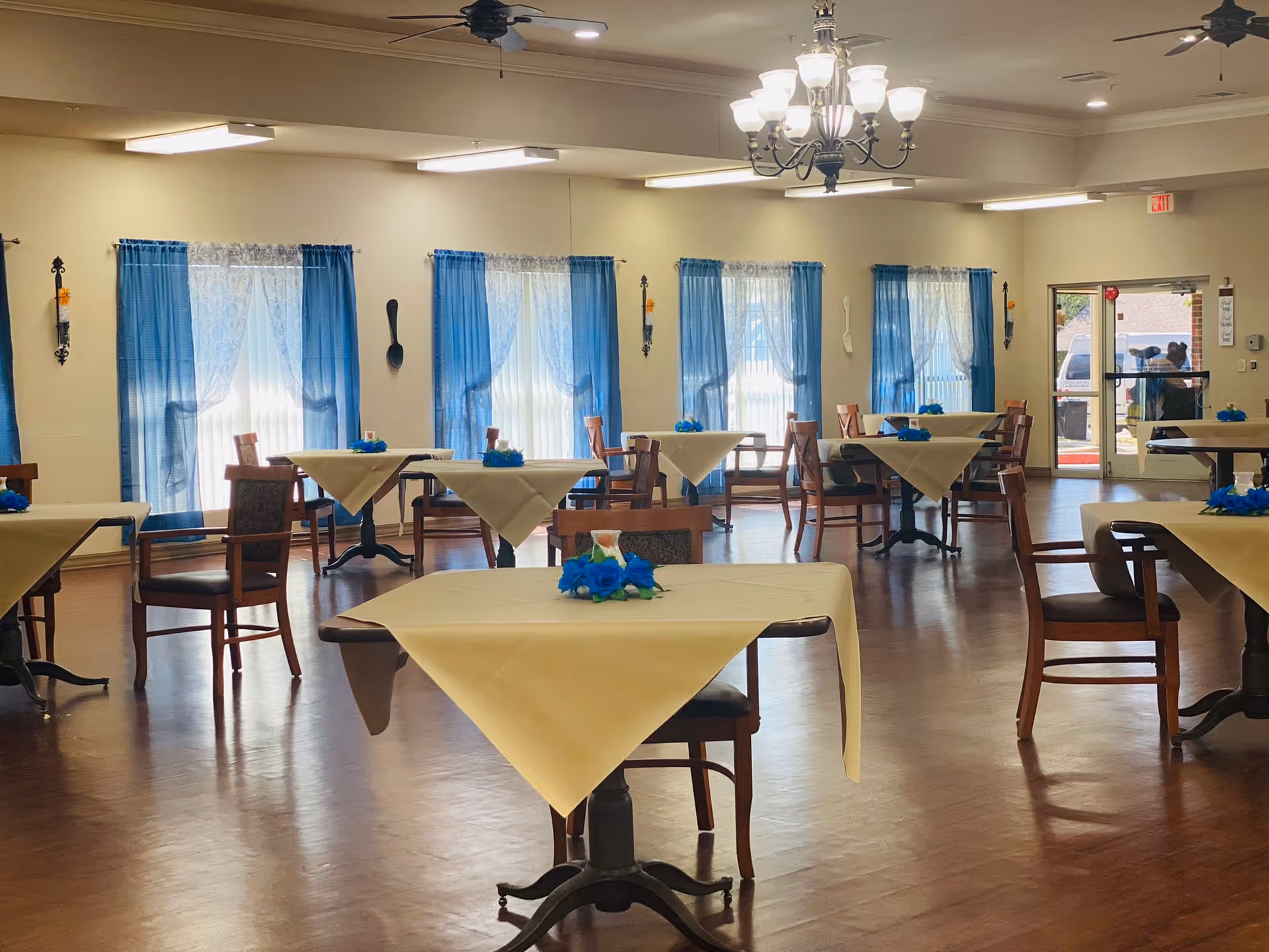 A spacious dining room with multiple square tables covered with beige tablecloths, each adorned with a small blue floral centerpiece. Wooden chairs are arranged around the tables. The room has large windows with blue curtains and white lace sheers, and the floor is wooden. Ceiling lights and a chandelier provide illumination. A glass door is visible at the far end of the room.