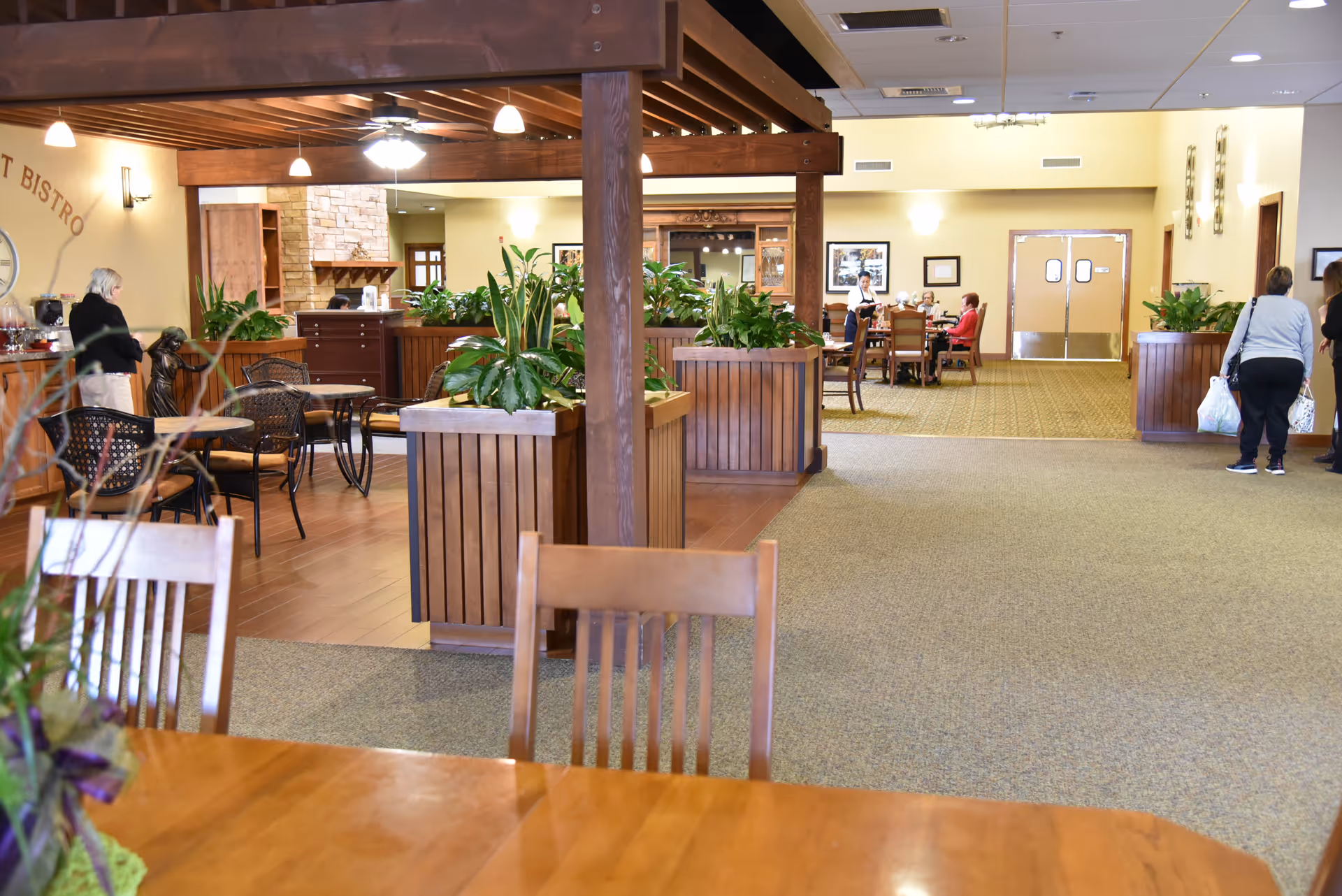Interior view of a senior living facility dining and common area with wooden tables and chairs, large planters with green plants, and people sitting and standing in the background. The space has warm lighting and a mix of carpet and wood flooring.