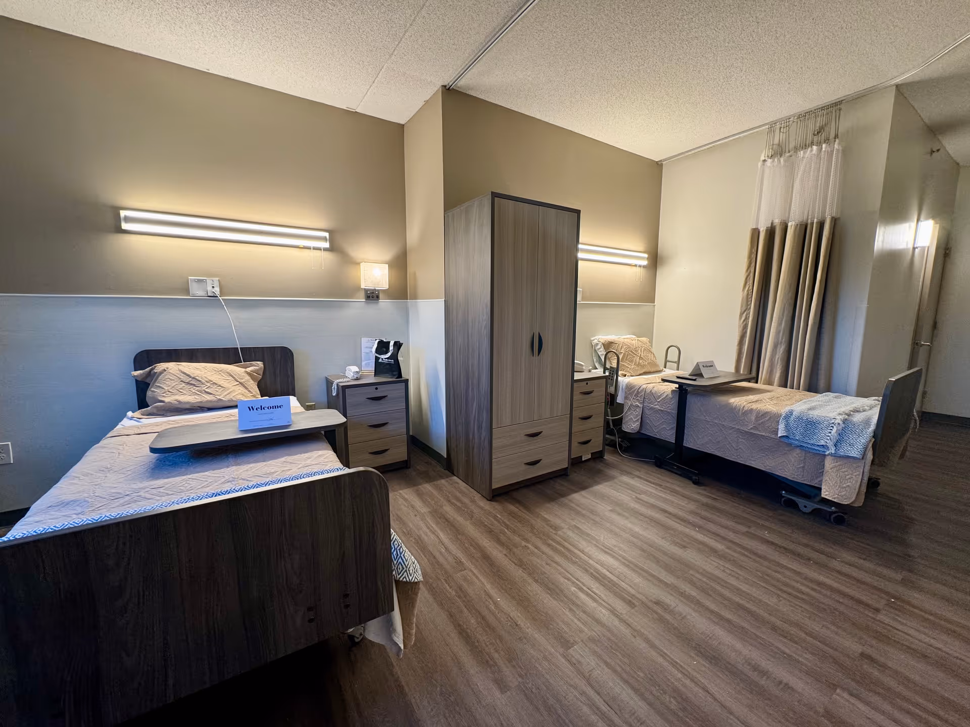 Shared nursing facility bedroom with two hospital-style beds, bedside tables, a central wardrobe, and privacy curtains.