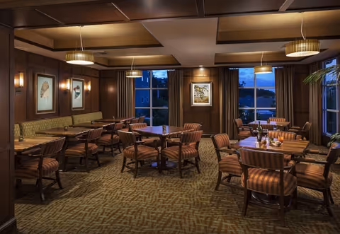 A warmly lit dining room with multiple wooden tables and striped cushioned chairs arranged neatly. The room features wood-paneled walls adorned with framed artwork, large windows with curtains showing an evening sky outside, and modern pendant lights hanging from the ceiling.