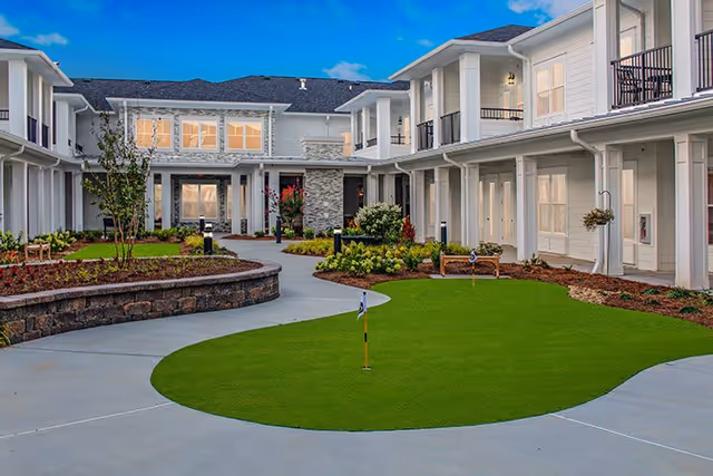 Outdoor courtyard area of The Blake at Kingsport featuring a small putting green, landscaped garden beds, benches, and a two-story white building surrounding the courtyard under a blue sky.