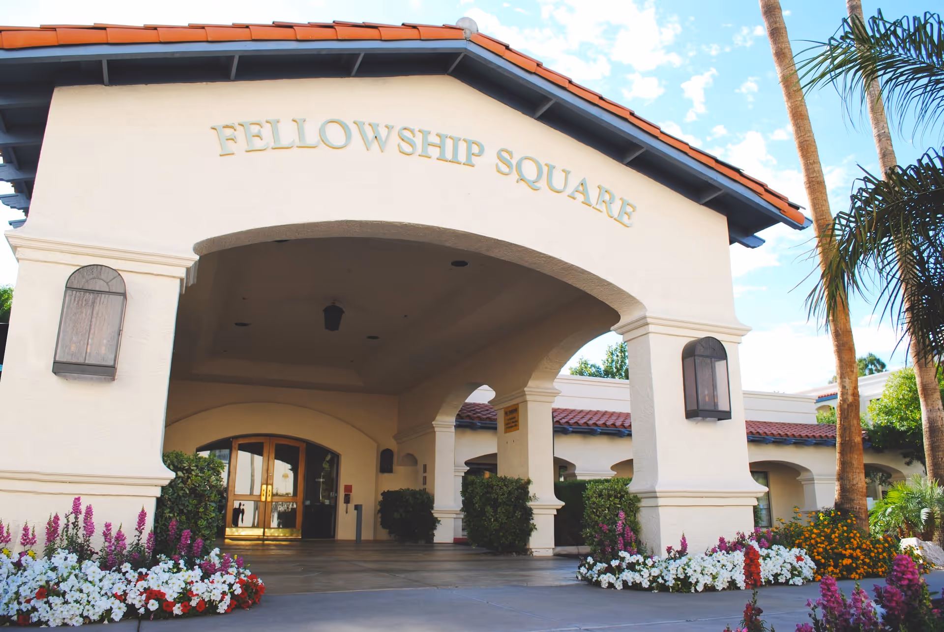 Entrance of Fellowship Square Phoenix facility with an archway featuring the name 'FELLOWSHIP SQUARE' above. The building has a tiled roof and is surrounded by colorful flower beds and palm trees under a partly cloudy sky.