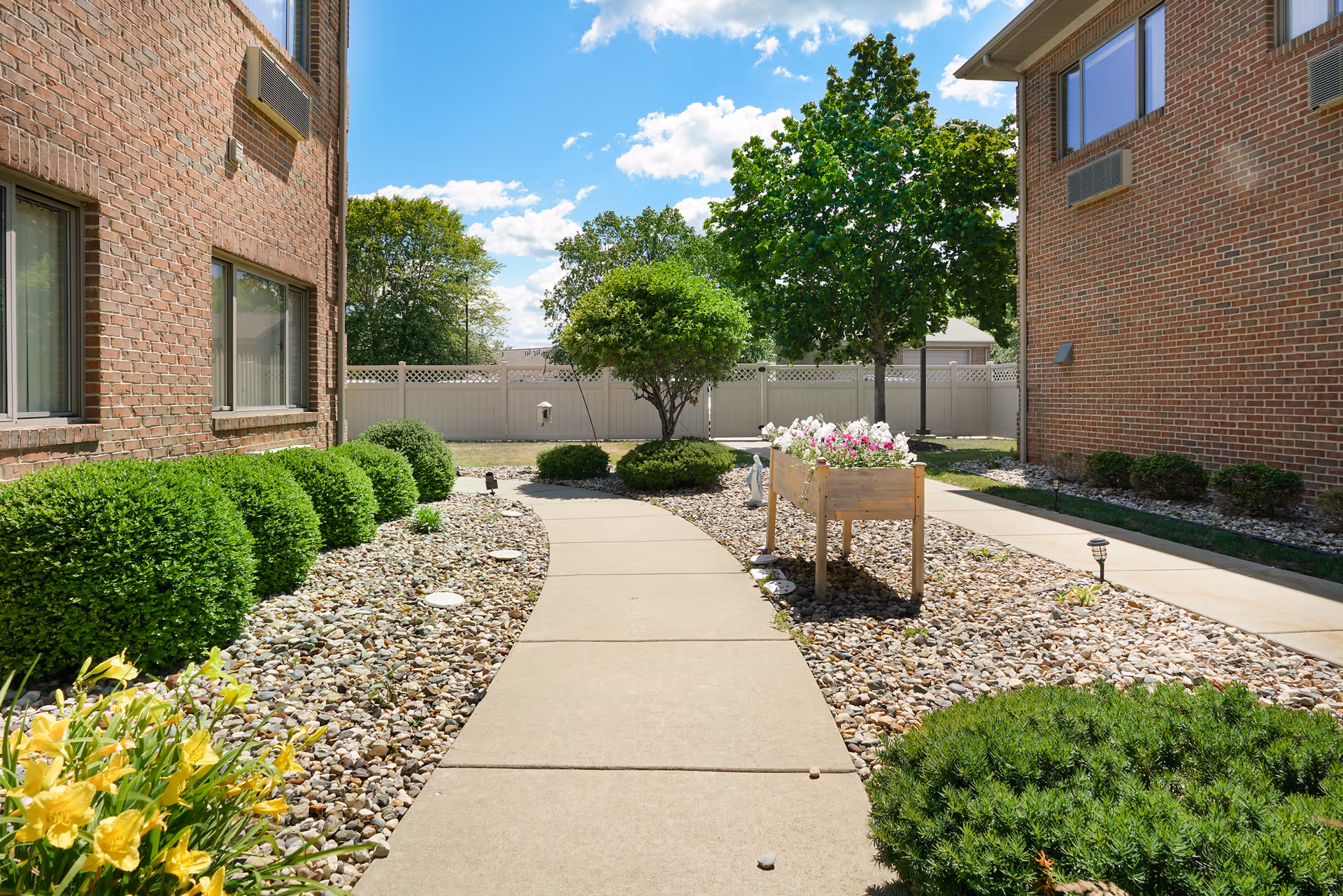A sunny outdoor walkway between two brick buildings with neatly trimmed bushes, a flower bed with blooming flowers, and trees in the background under a blue sky with scattered clouds.
