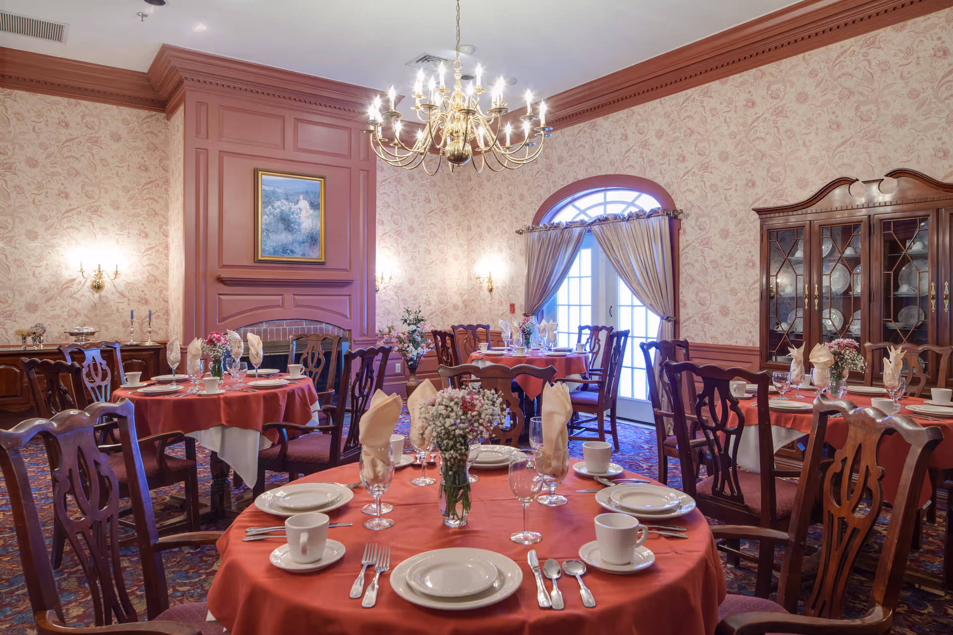 Elegant dining room with round tables covered in red tablecloths and set with white plates, cups, silverware, and folded napkins. The room features floral wallpaper, a chandelier, wall sconces, a fireplace with a framed painting above it, a glass-front china cabinet, and a door with curtains letting in natural light.