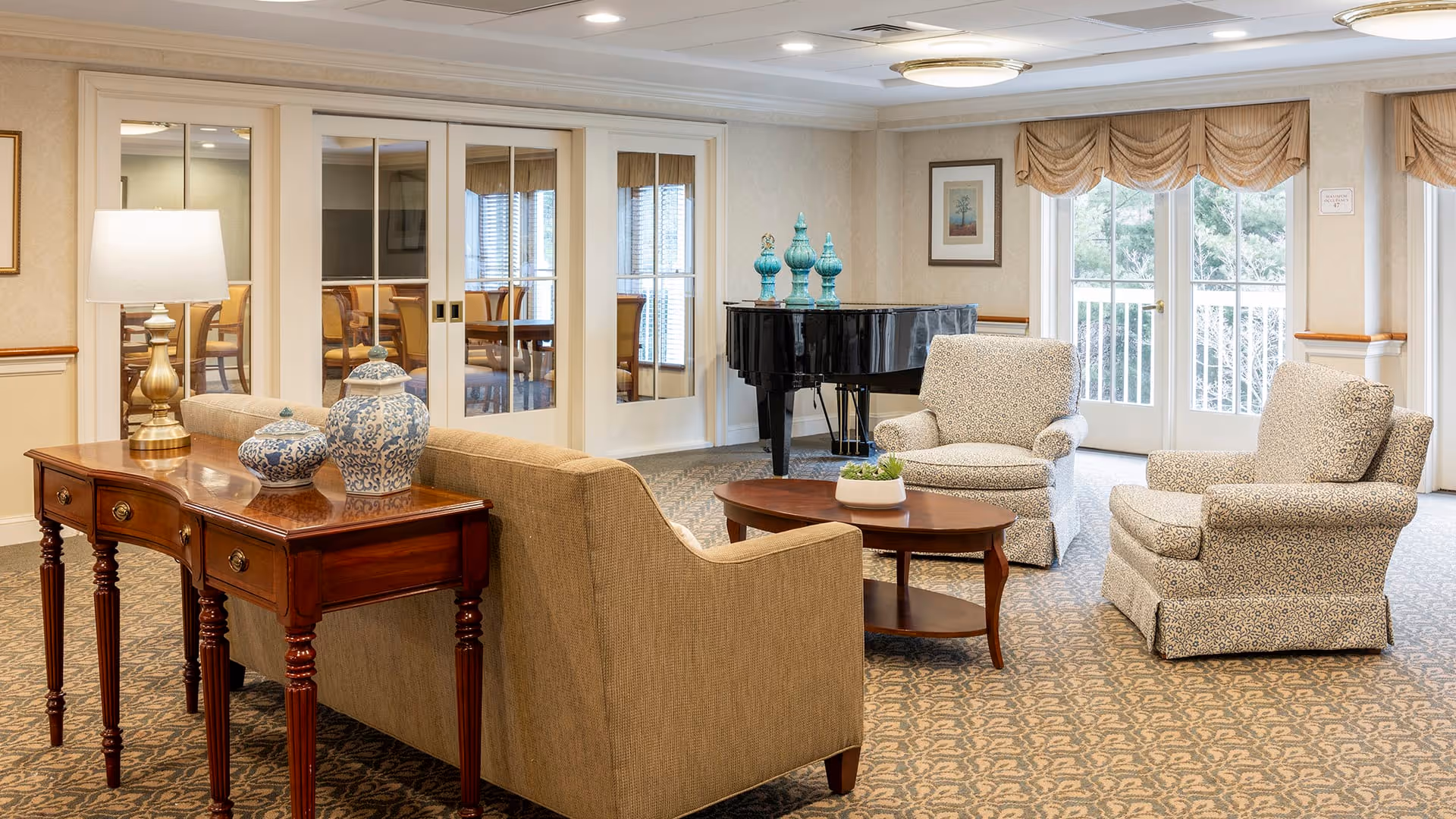 A cozy living room area with a beige sofa and two patterned armchairs arranged around a wooden coffee table with a small plant. Behind the sofa is a wooden console table with decorative blue and white vases and a lamp. In the background, there is a black grand piano with blue decorative items on top, glass-paneled sliding doors, and large windows with beige valances letting in natural light.