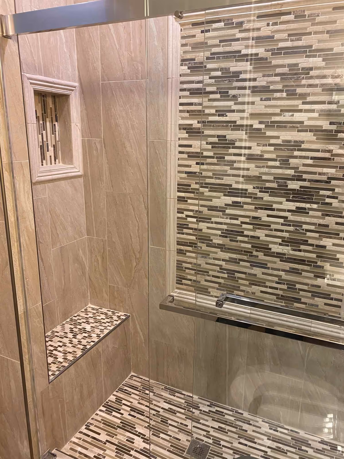 A modern shower area with beige and brown mosaic tiles on the floor and one wall, beige large tiles on the other walls, a built-in niche with mosaic tile accents, and a glass shower door with a metal handle.