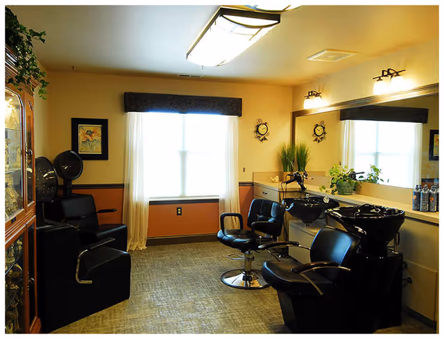 A small salon-style room with hair-wash sinks, styling chairs, a large mirror and a bright window.