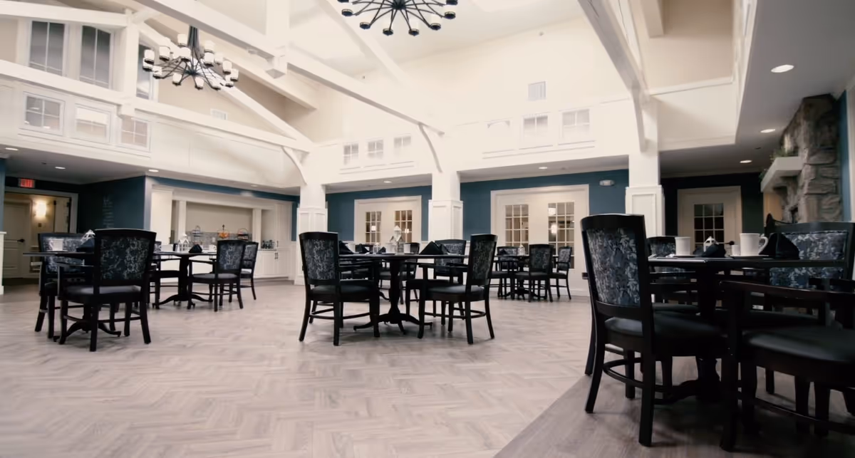 A spacious dining room with multiple round tables and chairs arranged neatly. The room features high vaulted ceilings with exposed beams and modern chandeliers. The walls are painted in a combination of white and blue, and there are large windows and doors along the walls. The floor has a light wood pattern, and the tables are set with cups and napkins.