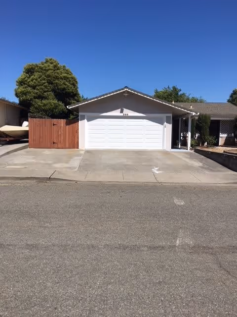 Front exterior view of a single-story house with a white garage door, a wooden fence on the left, and a clear blue sky above.
