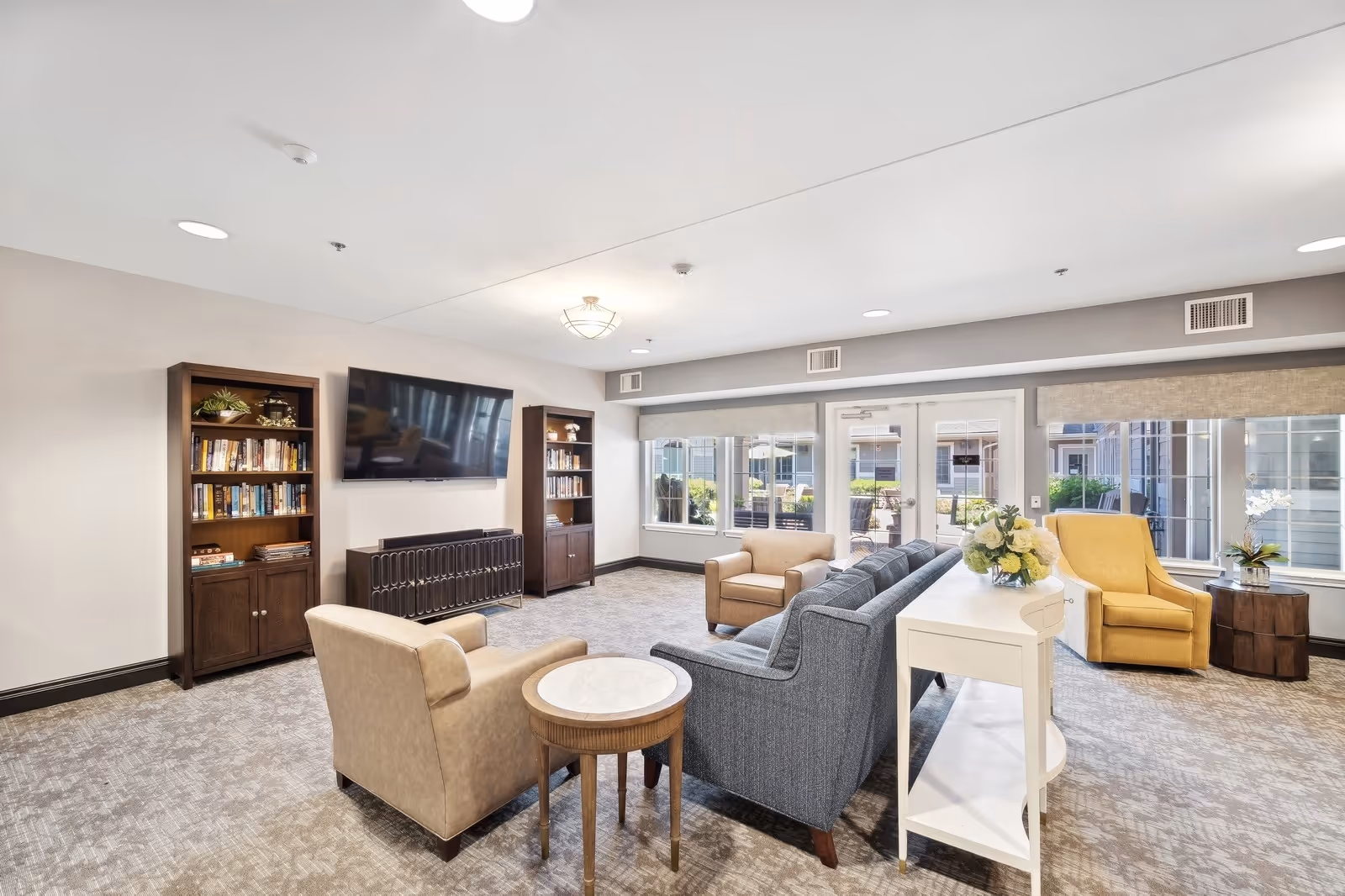 A bright and spacious living room area in a senior living facility featuring a gray sofa, beige armchairs, a yellow armchair, two wooden bookshelves filled with books, a wall-mounted flat-screen TV, and large windows with views of an outdoor patio area. The room is carpeted and decorated with a white console table holding a flower arrangement.