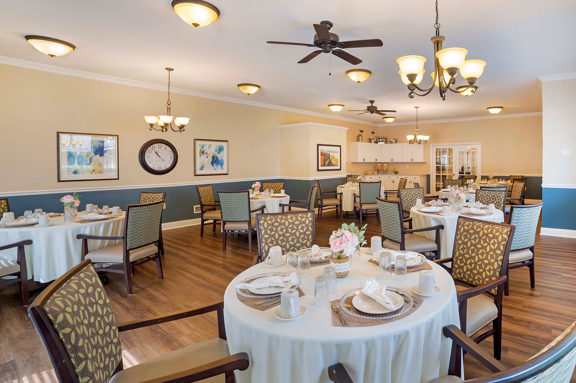 A bright and spacious dining room with round tables covered in white tablecloths, each set with plates, cups, napkins, and glasses. The room features wooden flooring, patterned chairs, ceiling fans, chandeliers, and framed artwork on the walls. There is a clock on one wall and a cabinet area in the background.