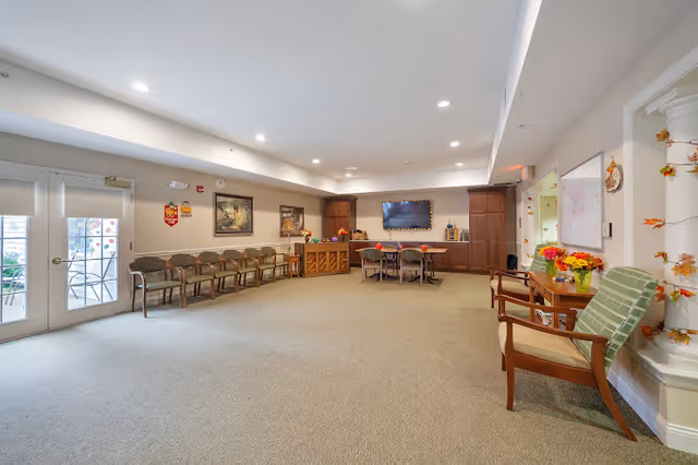 A spacious common area in a senior living facility with beige carpet and walls. There are several chairs lined up against the left wall near glass doors, a piano with decorations, a table with chairs, and a large flat-screen TV mounted on the far wall. On the right side, there are two wooden chairs with cushions and a small table with a vase of flowers. The room is well-lit with recessed ceiling lights and decorated with autumn-themed items.