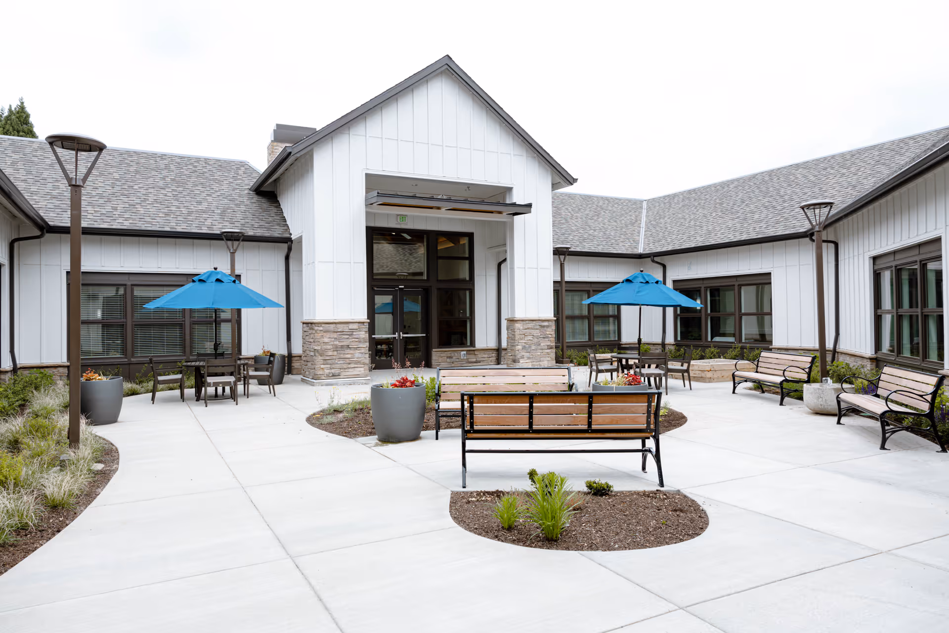 Outdoor courtyard area of The Springs at Happy Valley featuring benches, tables with blue umbrellas, potted plants, and modern building exterior with large windows and stone accents.