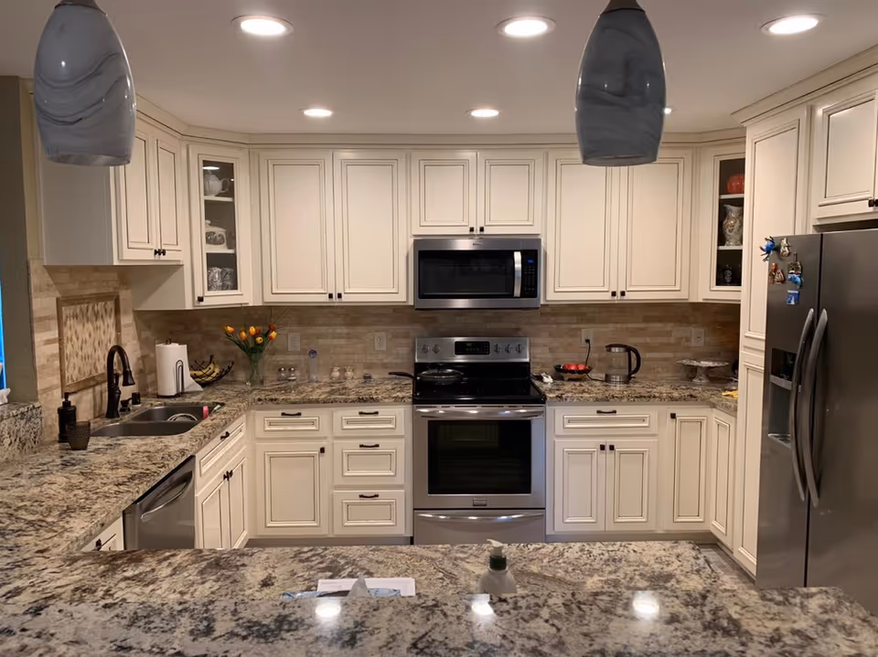 A modern kitchen with cream-colored cabinets, granite countertops, and stainless steel appliances including a microwave, stove, dishwasher, and refrigerator. The kitchen features a double sink, pendant lights, and decorative items such as a vase with flowers and a bowl of fruit.