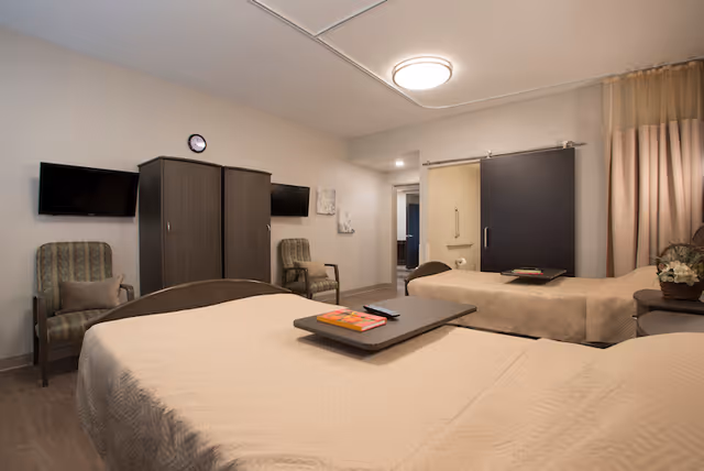 A shared bedroom with two neatly made beds, armchairs, wardrobes, wall-mounted TVs, and a sliding bathroom door.