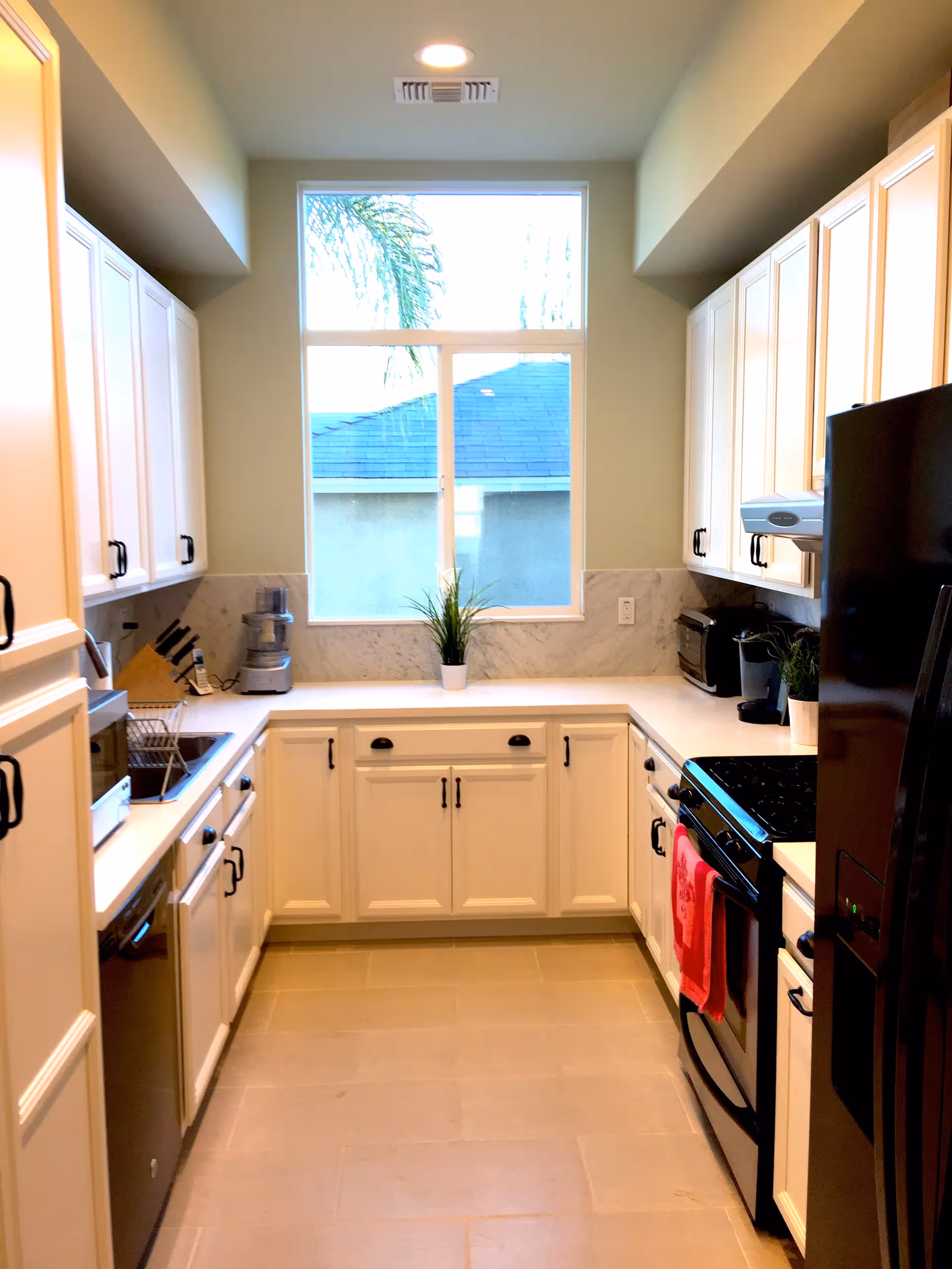 A bright, modern kitchen with white cabinets and black handles, a large window letting in natural light, a black refrigerator, stove with a red towel hanging on the handle, a dishwasher, a toaster, a coffee maker, a food processor, and a small potted plant on the countertop.