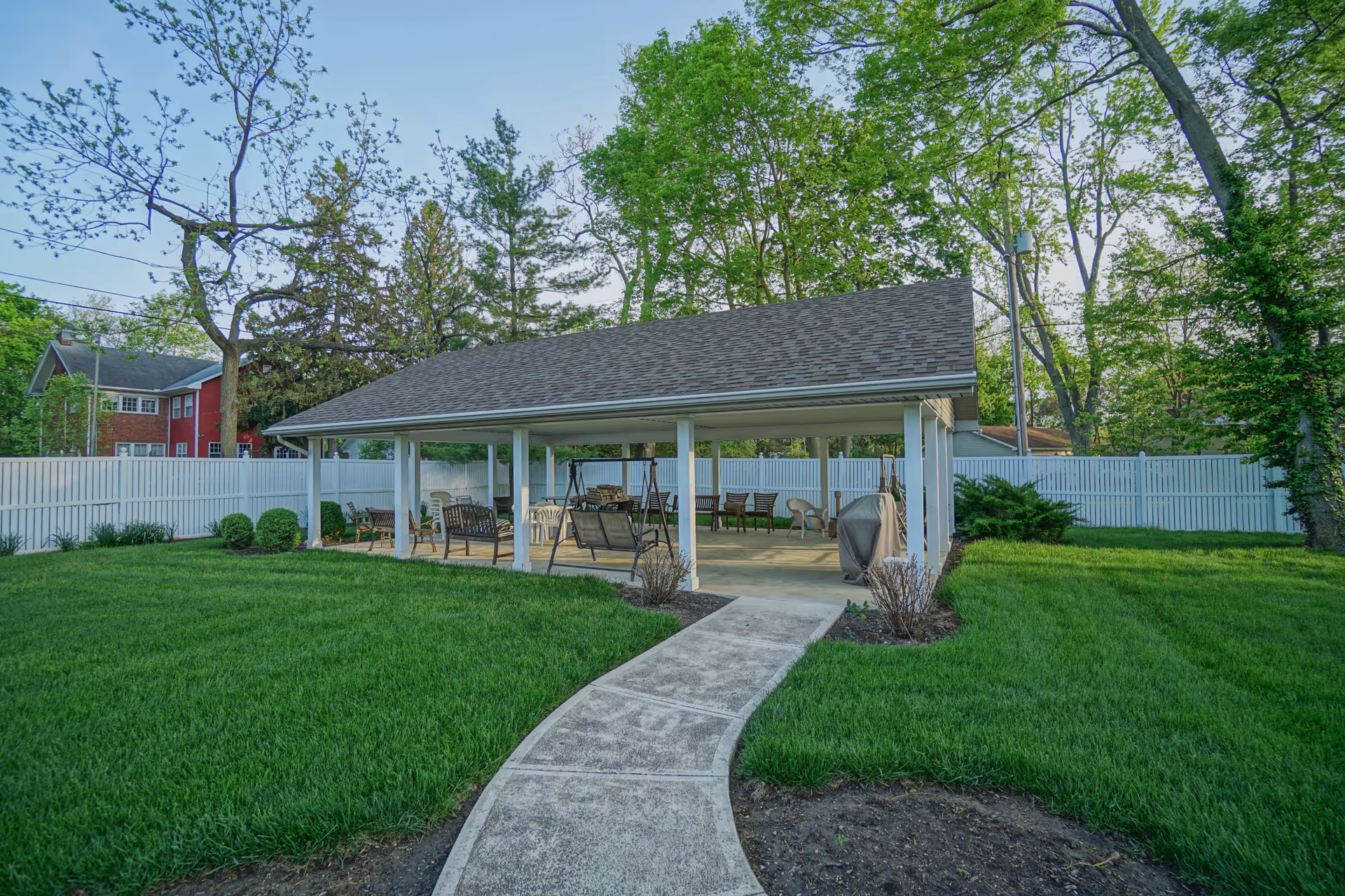 A covered outdoor pavilion with seating and swings in a grassy yard surrounded by a white fence and trees.