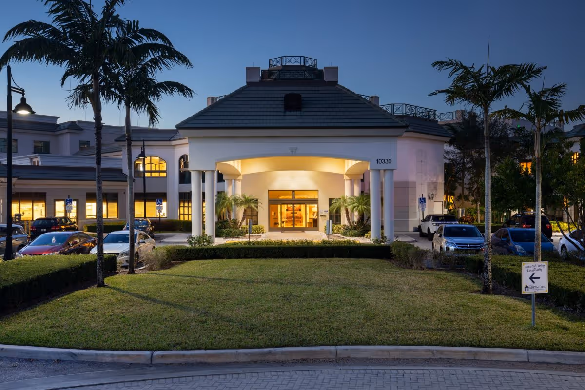 Entrance of a senior living facility at dusk showing a lit porte-cochère, palm trees, lawn, and parked cars.