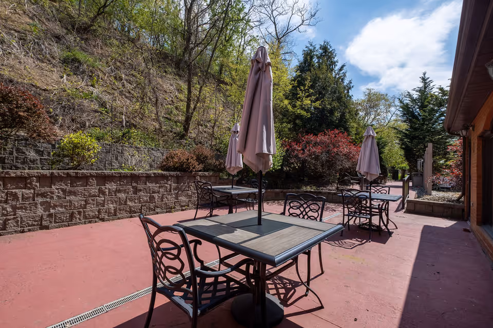Outdoor patio with metal tables, chairs, and closed umbrellas next to a retaining wall and wooded hillside.
