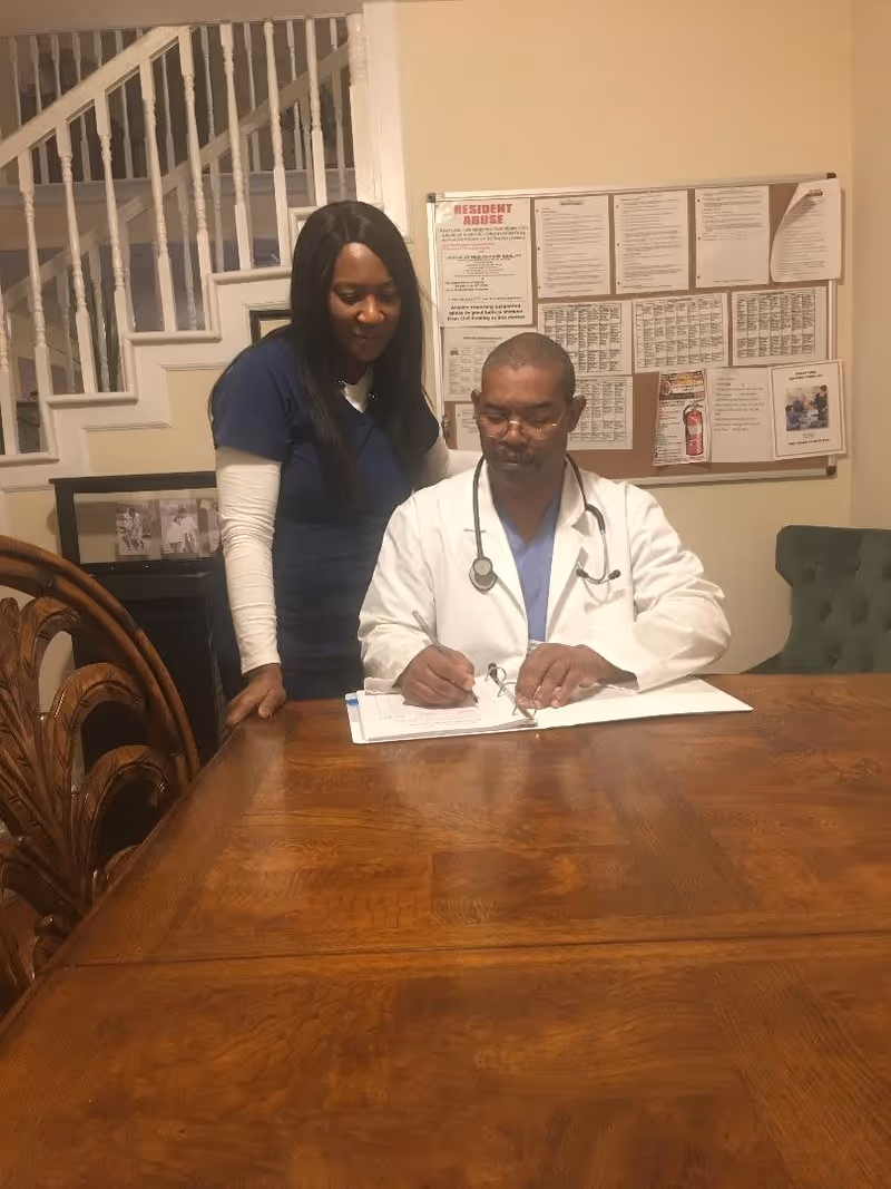 A man in a white medical coat with a stethoscope around his neck is sitting at a wooden table writing in a binder, while a woman in navy blue scrubs stands beside him looking at the binder. Behind them is a bulletin board with various notices and a staircase with white railings.