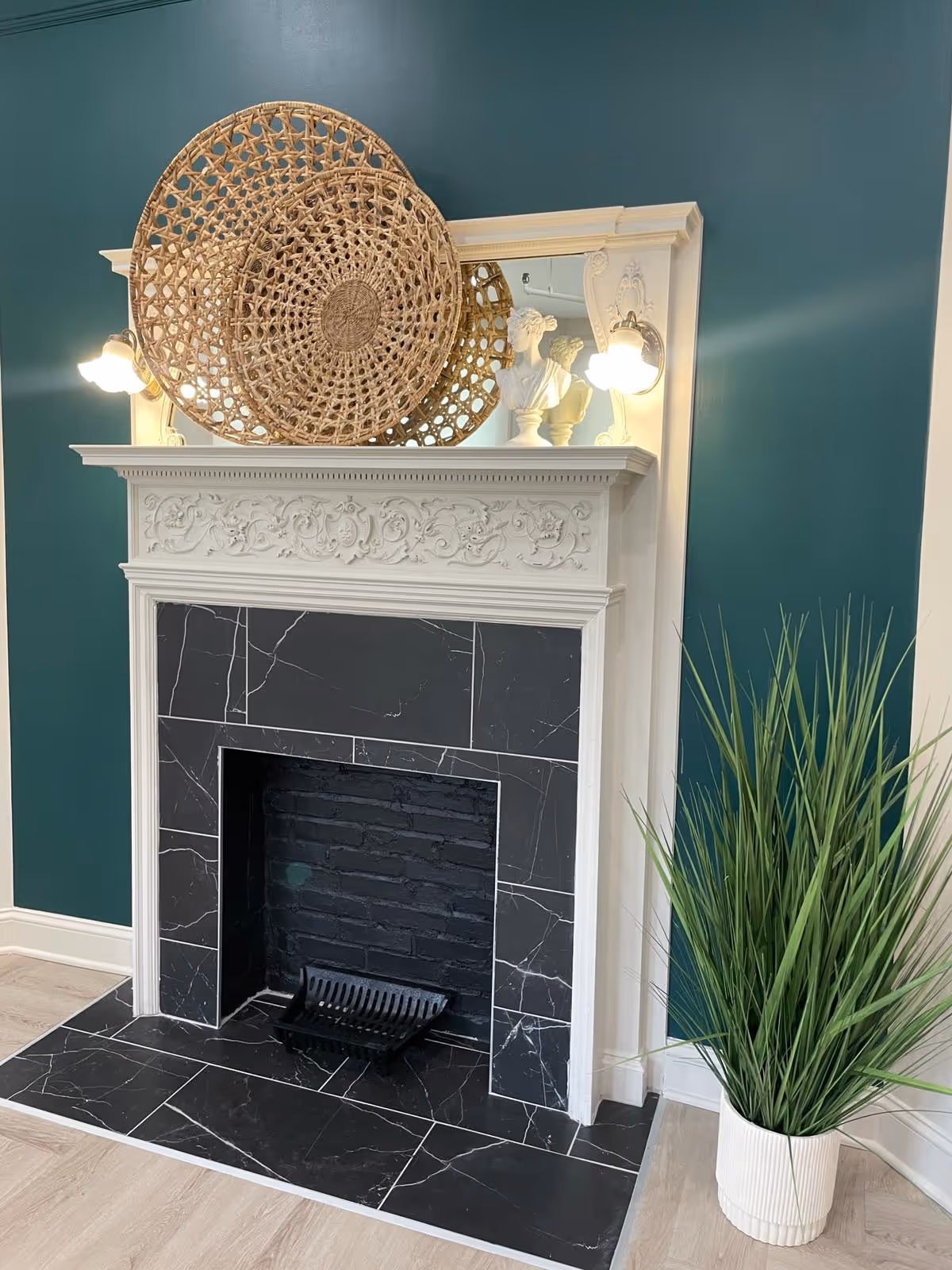 A decorative fireplace with a white ornate mantel and black marble tiles surrounding the firebox. On top of the mantel are two woven circular baskets and a white bust sculpture, flanked by two wall-mounted light fixtures. To the right of the fireplace is a tall green plant in a white pot. The wall behind is painted teal.