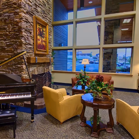 A cozy living room area with a stone fireplace, a grand piano, yellow armchairs, a wooden coffee table with plants, and large windows showing an outdoor view.