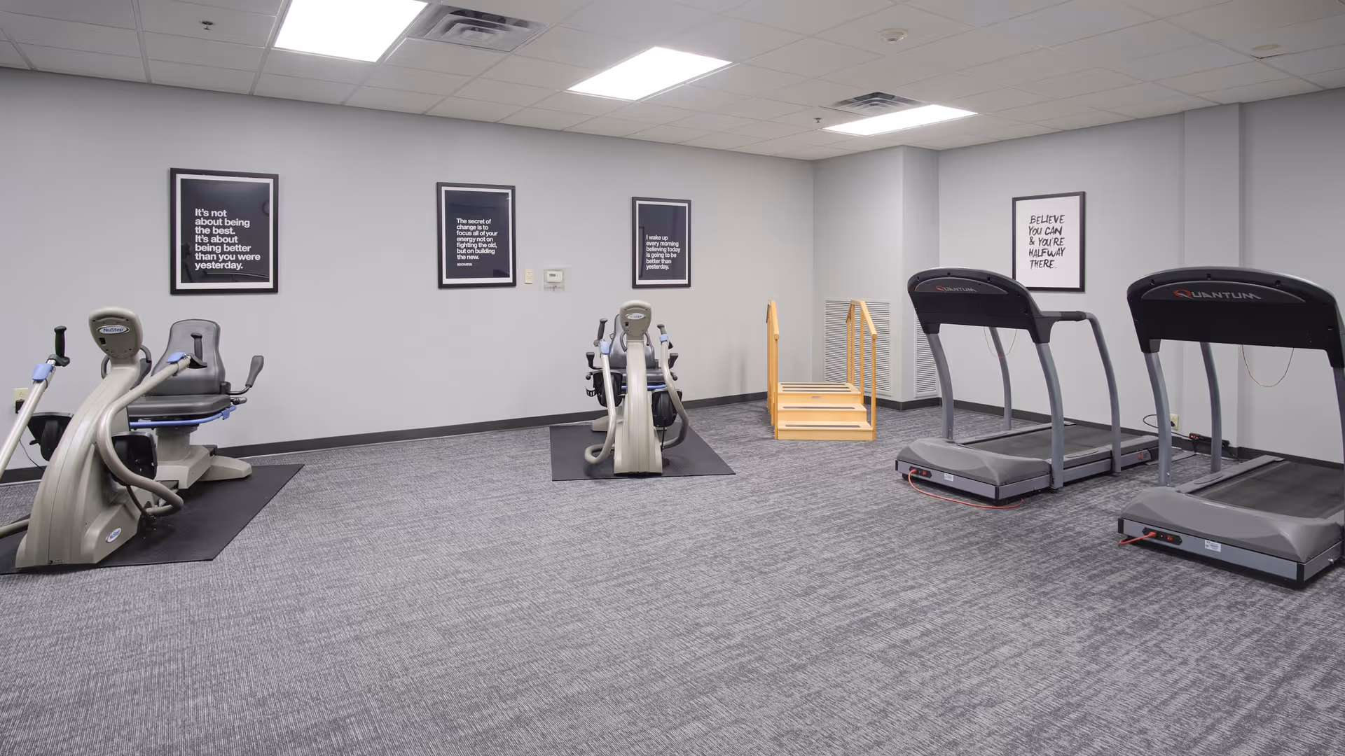 A senior fitness room with treadmills, recumbent exercise bikes, and framed motivational posters on gray walls.