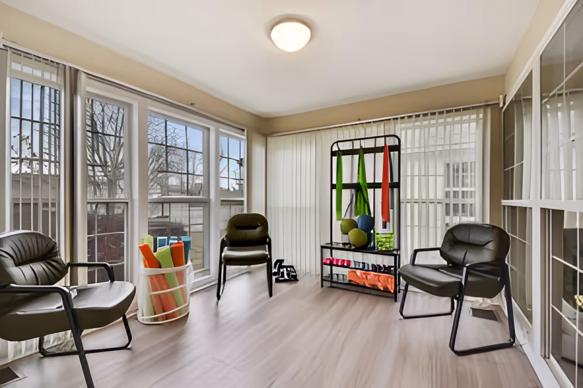 A bright room with large windows on two sides, featuring three black chairs arranged around the space. There is a metal rack holding various colorful exercise bands, balls, and small weights. A container with rolled-up yoga mats is placed near the windows. The room has light-colored wooden flooring and vertical blinds covering the windows.