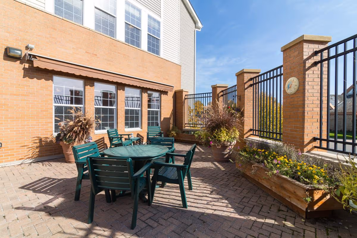 Sunny brick patio with a green plastic table and chairs, potted plants, and a fenced brick wall under windows.