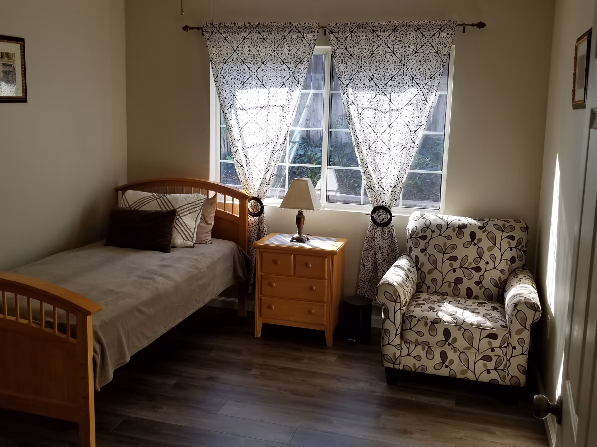 A small bedroom with a single wooden bed covered with a beige blanket and three pillows. Next to the bed is a wooden nightstand with a lamp on it. A window with patterned white curtains lets in natural light. To the right of the nightstand is a cushioned armchair with a floral pattern. The room has wooden flooring and light-colored walls.