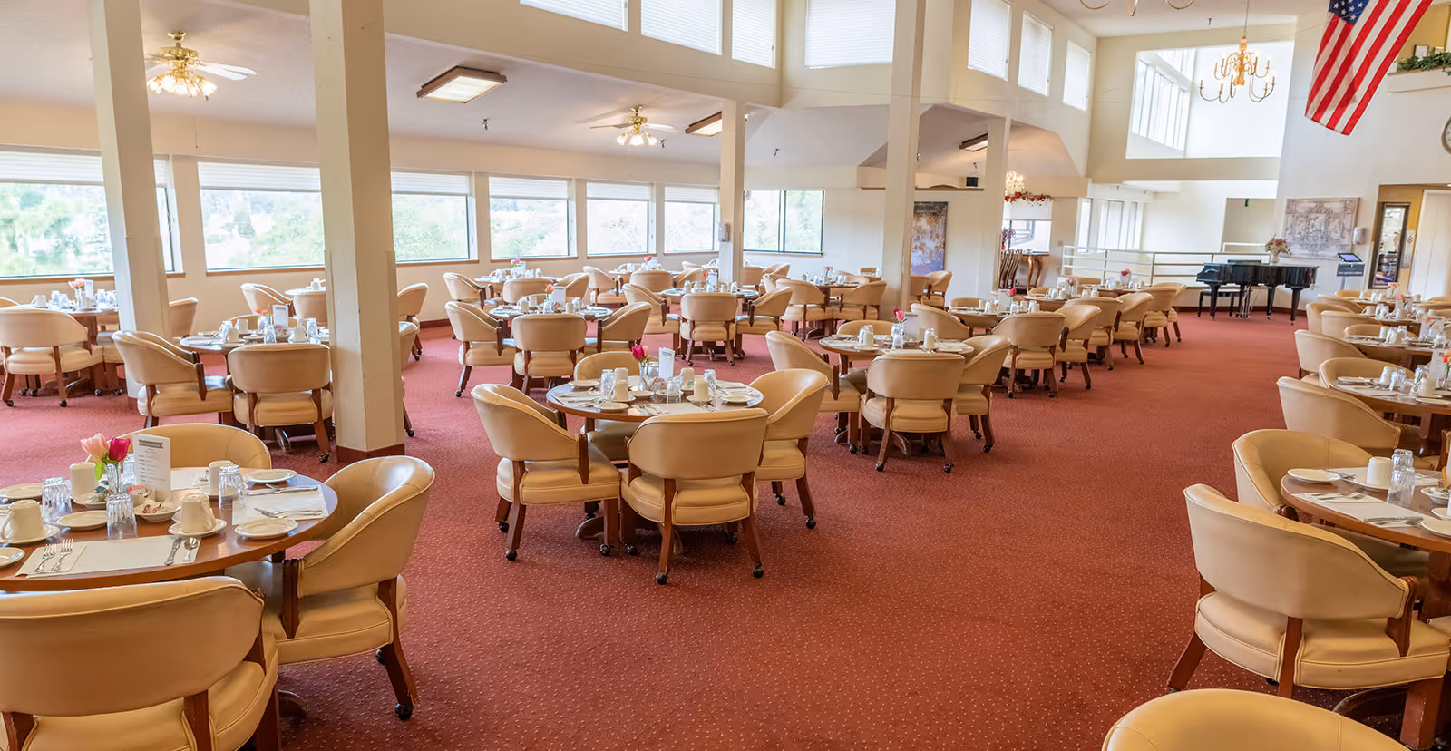 Spacious dining room with multiple round tables set with plates, cups, and silverware. Beige cushioned chairs surround each table. Large windows allow natural light to fill the room, and an American flag hangs from the ceiling. A grand piano is visible in the background.