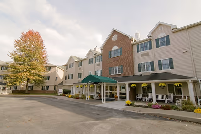 Exterior view of a multi-story senior living facility with beige siding and a brick central section. The entrance features a green awning and white columns, with rocking chairs and hanging flower baskets on the porch. A large tree with autumn-colored leaves is visible on the left side of the building.