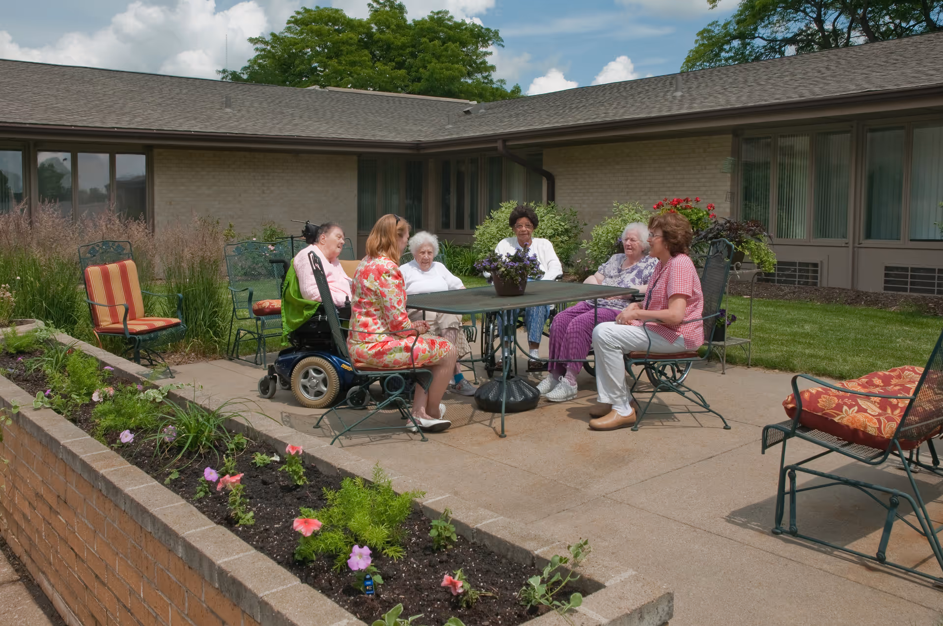 A group of elderly women sitting and chatting around a metal outdoor table with a flower pot centerpiece on a patio. The patio is part of a single-story brick building with large windows. There are additional empty chairs with cushions and a flower bed with blooming flowers in the foreground. The sky is partly cloudy.