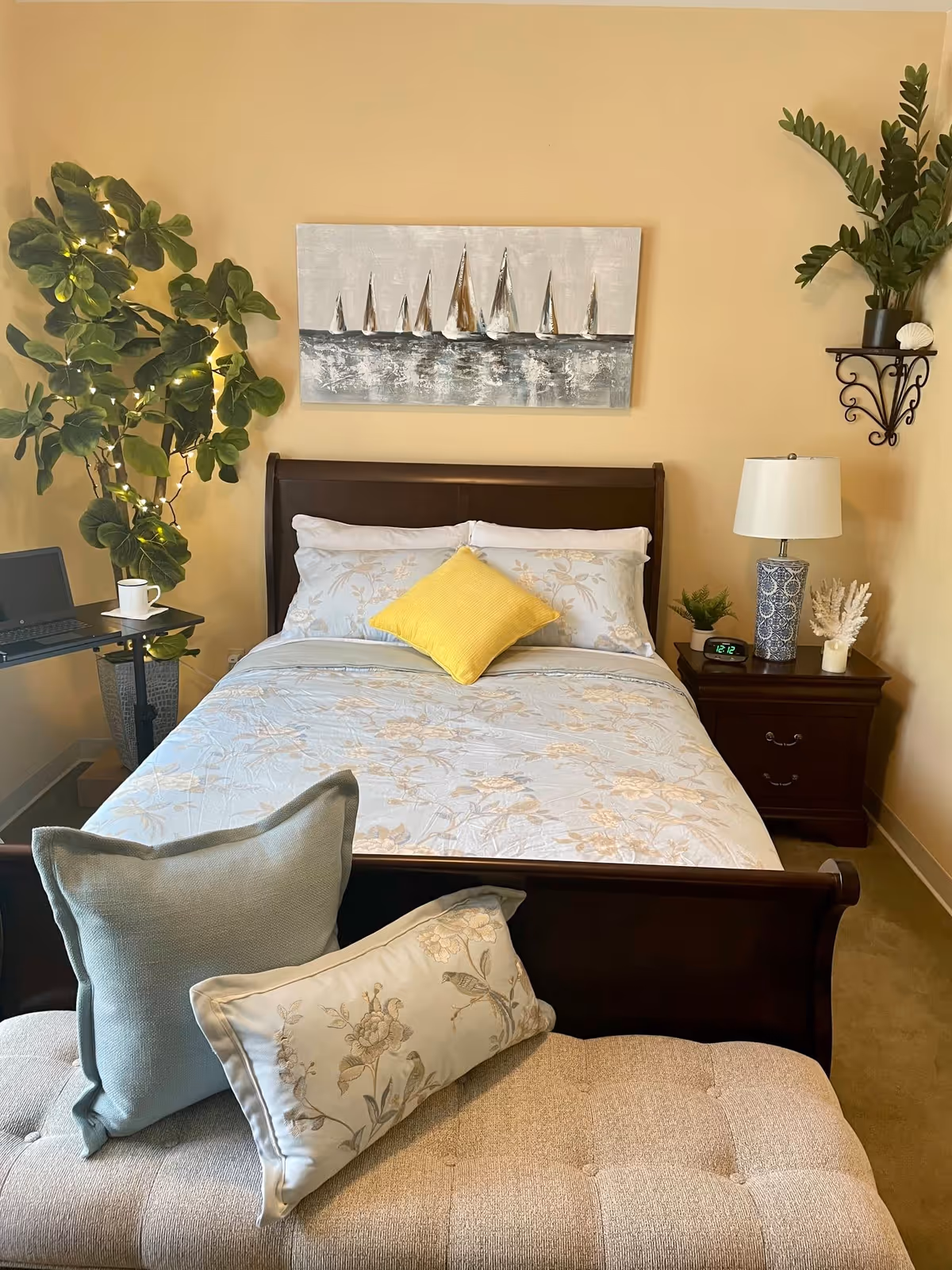 A cozy bedroom with a dark wooden bed frame, floral bedding, and a yellow accent pillow. There is a cushioned bench at the foot of the bed with two decorative pillows. On the left side, there is a small table with a laptop and a white mug, next to a large leafy plant with string lights. On the right side, a wooden nightstand holds a blue and white patterned lamp, a small plant, a clock showing 12:12, and a decorative coral piece. Above the bed hangs a painting of sailboats on water. The walls are painted a warm beige color.