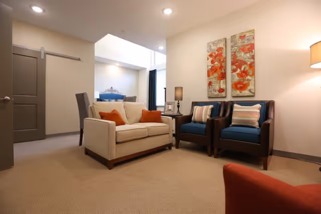 A cozy living room area with a beige loveseat featuring two orange pillows, two dark brown armchairs with blue cushions and striped pillows, a side table with a lamp and picture frame, and two floral paintings on the wall. The room has beige carpet and neutral-colored walls with recessed lighting.