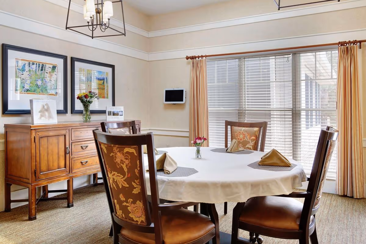 Bright dining room with a round table set with folded napkins, upholstered chairs, a wooden sideboard with artwork and flowers, and large windows with blinds.