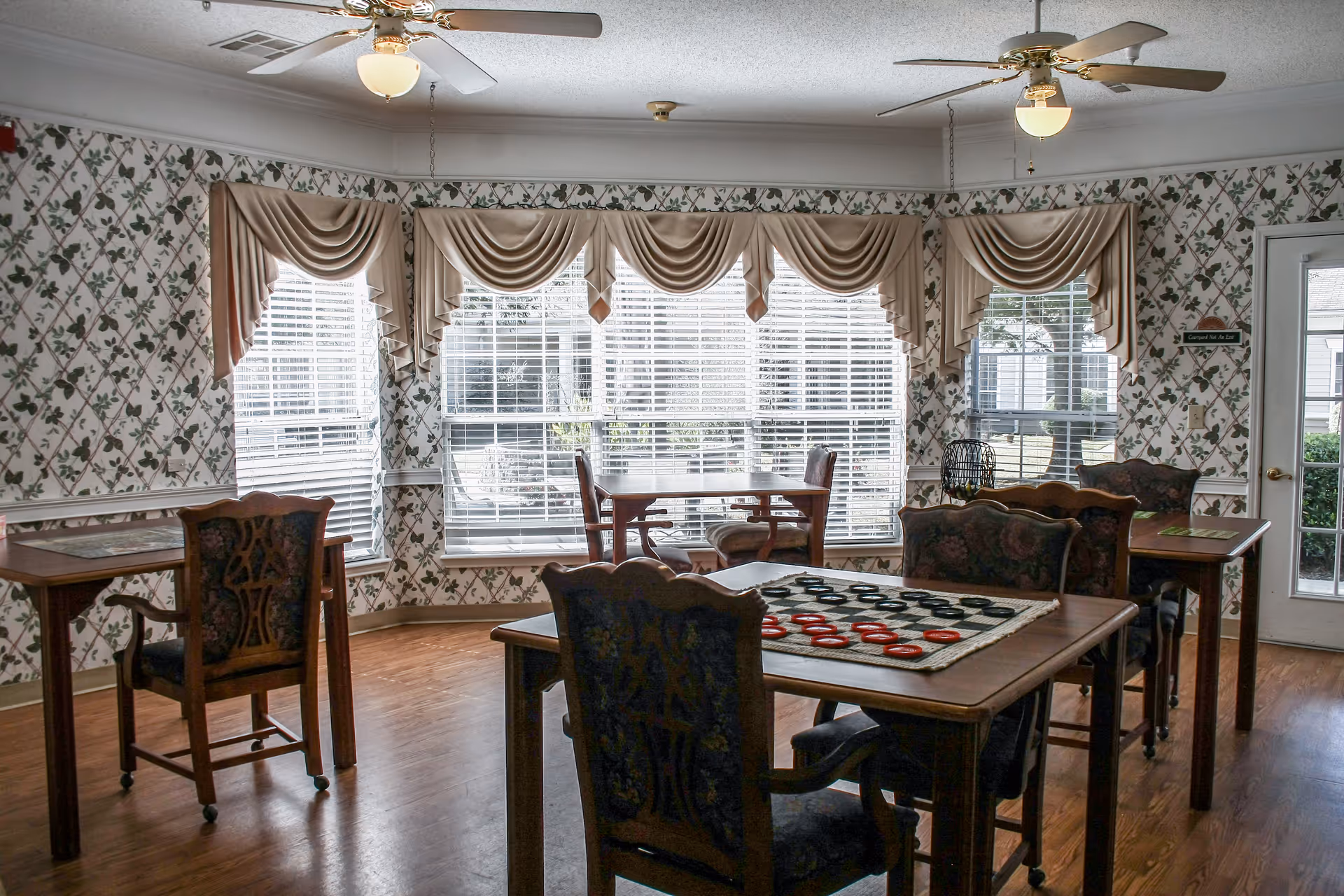 Sunlit common room with wooden tables and upholstered chairs, a checkerboard on a table, and large windows with draped valances.
