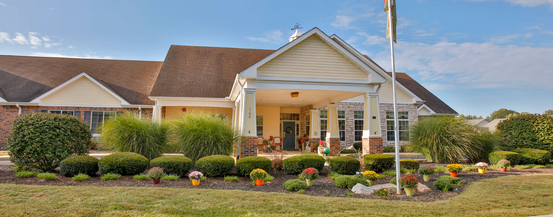 Front exterior view of a single-story senior living facility building with a covered entrance supported by columns. The building has a combination of brick and siding with a brown shingled roof. There are well-maintained bushes, ornamental grasses, and colorful potted flowers in the landscaped garden in front of the building. A flagpole stands near the entrance under a partly cloudy blue sky.