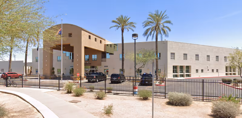 Exterior view of a large beige building with a covered entrance supported by columns, several windows, and palm trees in front. There is a black metal fence enclosing a parking area with several parked vehicles. The sky is clear and blue.
