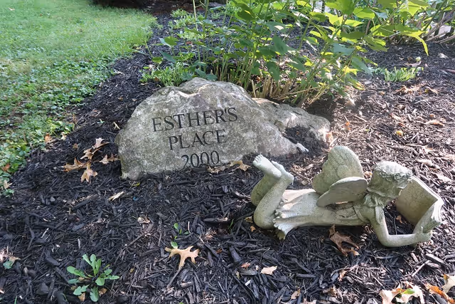 Garden bed showing a carved stone engraved "ESTHER'S PLACE 2000" beside a small reclining angel statue.