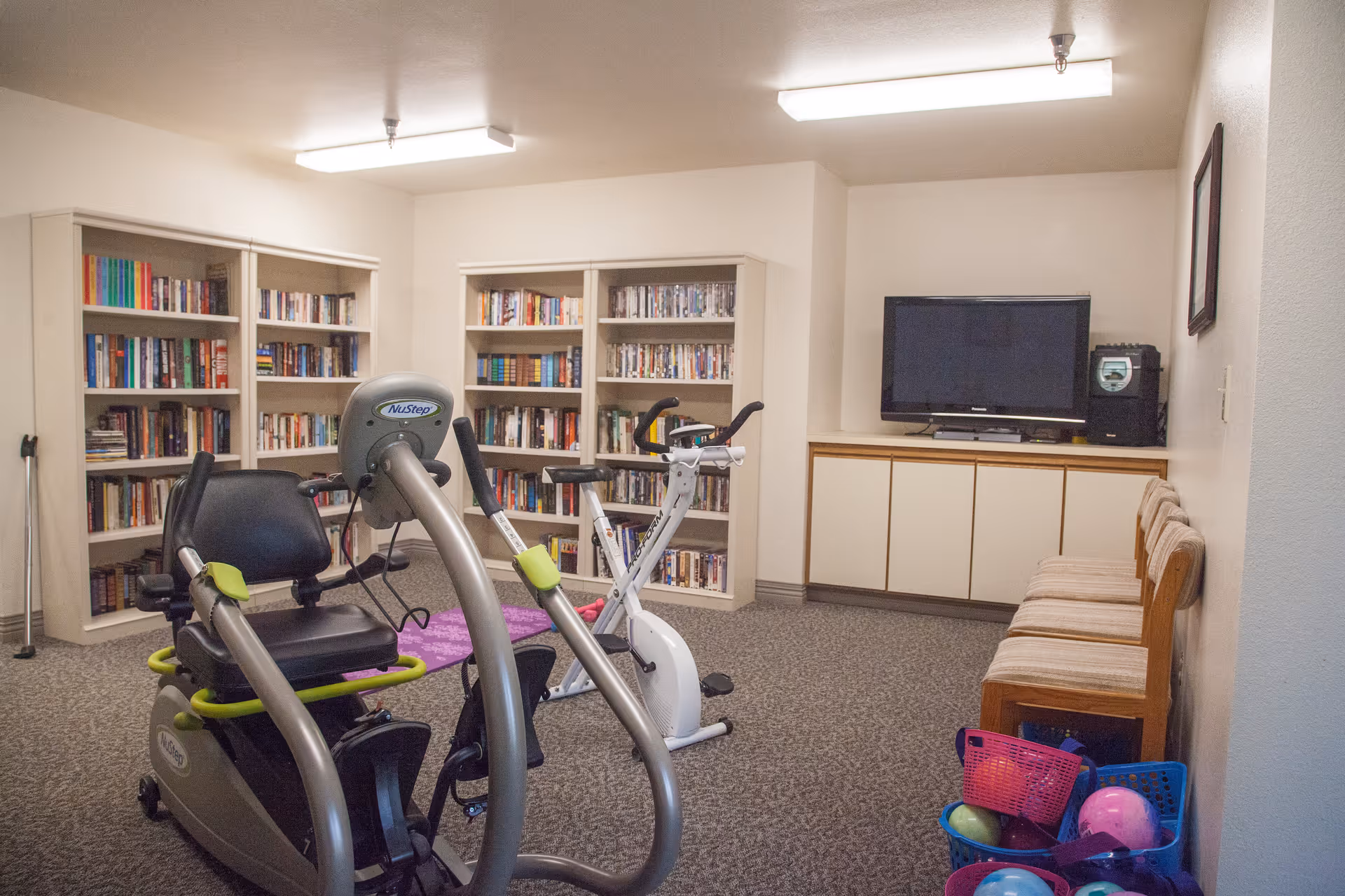 A small exercise room with two stationary exercise bikes in the center. Behind the bikes are bookshelves filled with books and DVDs. On the right side, there is a TV on a cabinet with three wooden chairs lined up against the wall. Several colorful plastic baskets containing balls are on the floor near the chairs. The room has carpeted flooring and fluorescent ceiling lights.