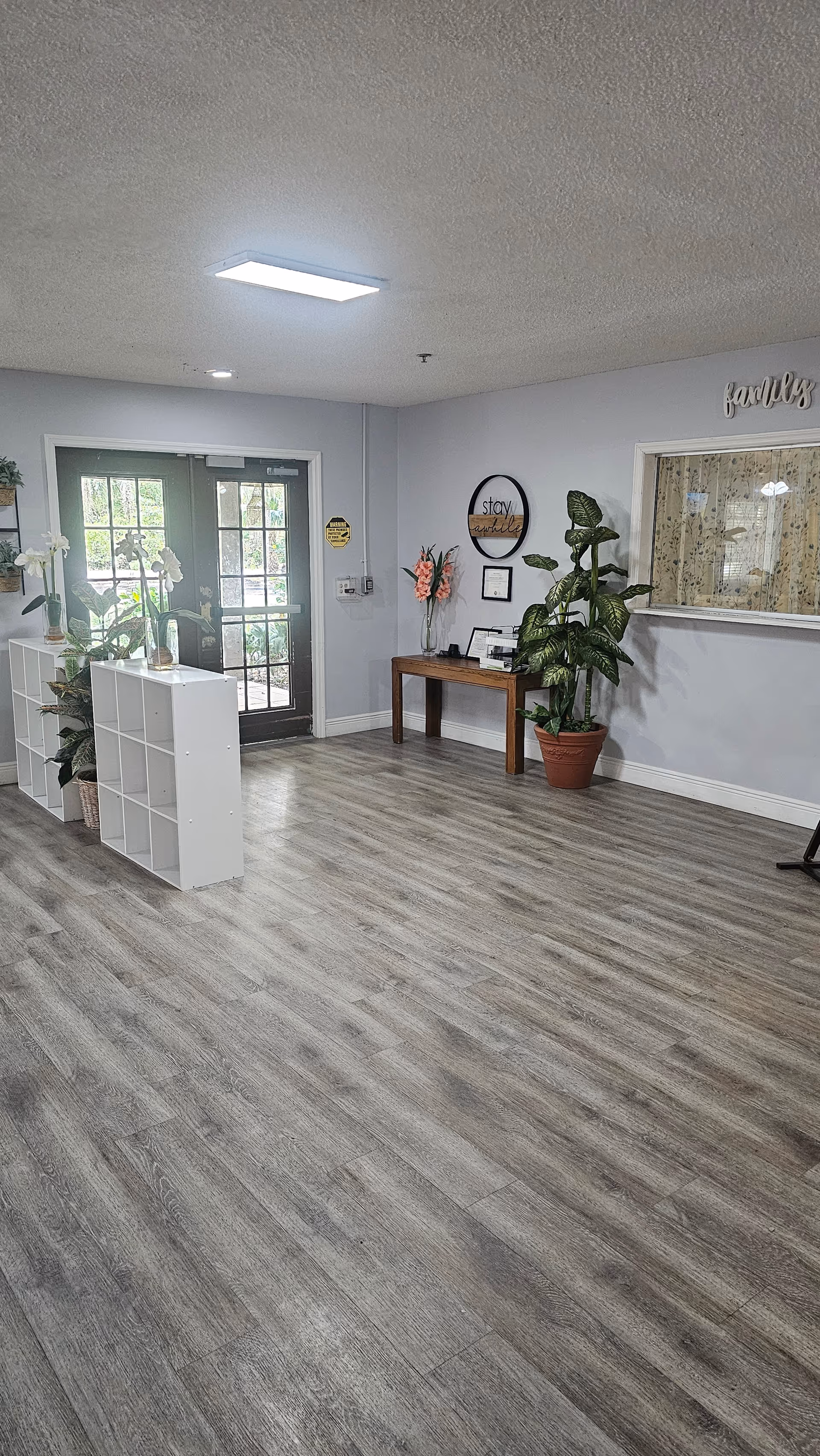 Interior view of a clean and bright assisted living facility lobby area with light gray walls and wood-look flooring. There are double glass doors letting in natural light, white shelving units with potted plants and flowers, a wooden table with decorative items and plants, and a window with a curtain on the right wall. The word 'family' is displayed above the window.