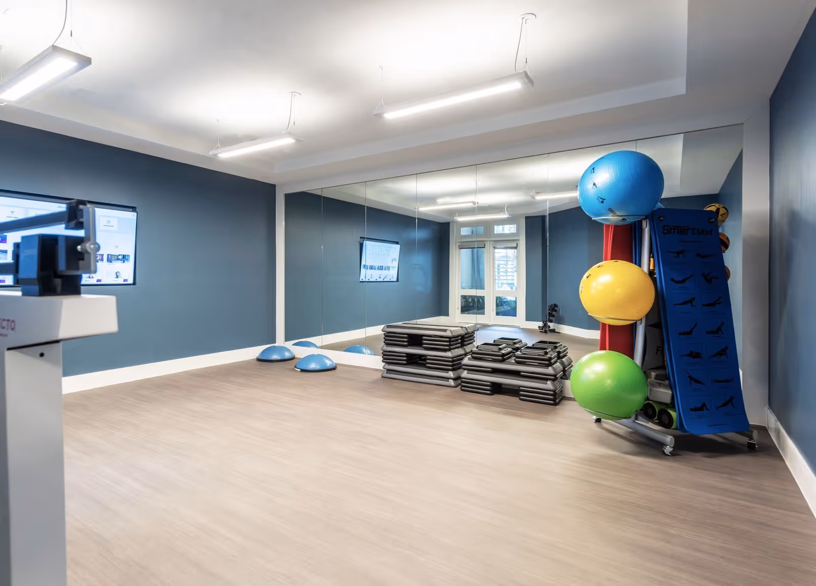 A bright mirrored fitness studio with stacked aerobic steps, colorful exercise balls, balance trainers, and a wall-mounted screen.