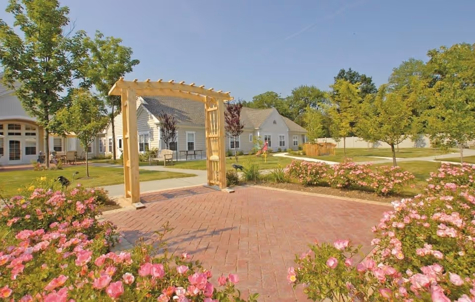 Outdoor garden area at Yorktown Assisted Living Residence featuring a wooden pergola archway, pink flowering bushes, green trees, paved walkways, and light-colored residential buildings in the background under a clear blue sky.