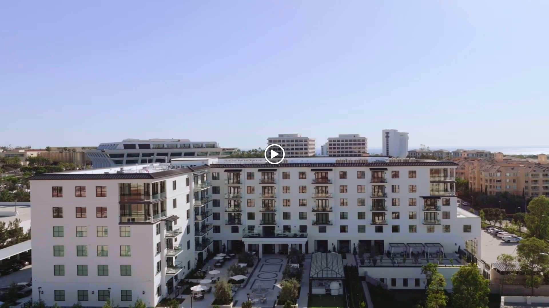 Aerial view of Vivante Newport Center, a multi-story senior living facility with balconies and a central courtyard featuring seating areas and landscaping. Surrounding buildings and a clear blue sky are visible in the background.