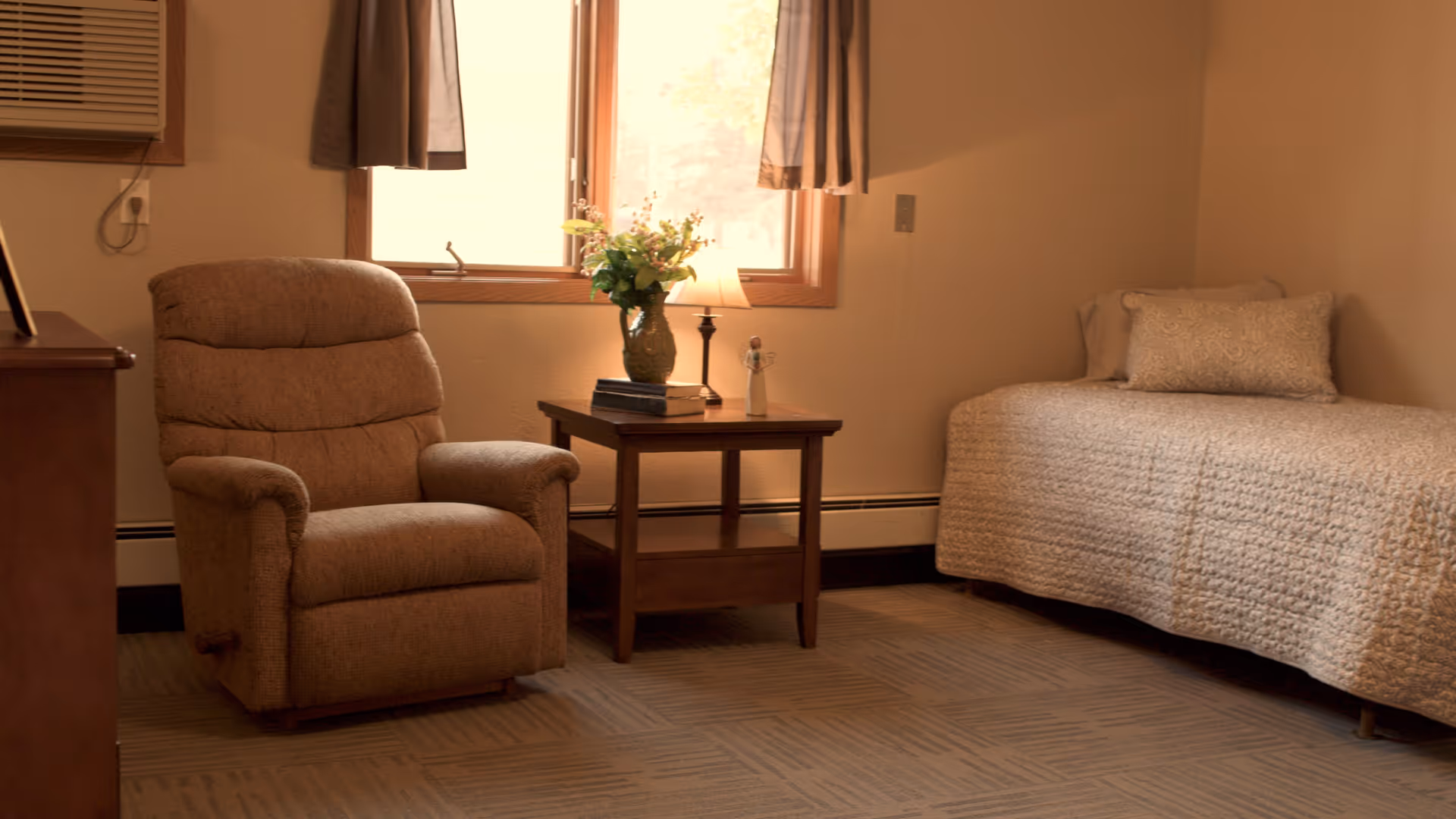 A cozy room with a beige recliner chair, a wooden side table with a vase of flowers and a small lamp, and a single bed with a textured beige bedspread and a decorative pillow. The room has a window with curtains and neutral-colored walls.